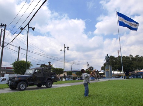 Un niño eleva una piscucha (cometa) en una plaza de San Salvador custodiada por un vehículo militar artillado.