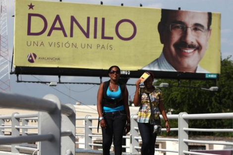 Dos mujeres caminan cerca de un cartel del candidato presidencial oficialista Danilo Medina. | Efe
