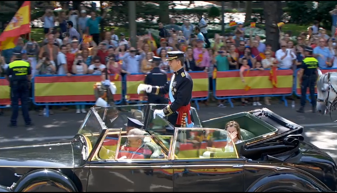 El Rey Felipe VI, de pie, y la Reina Letizia, sentada, saludando a los asistentes a su paso por el Paseo del Prado de Madrid.