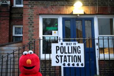 Una mujer disfrazada de Elmo espera a Jeremy Corbyn en la puerta del colegio electoral.