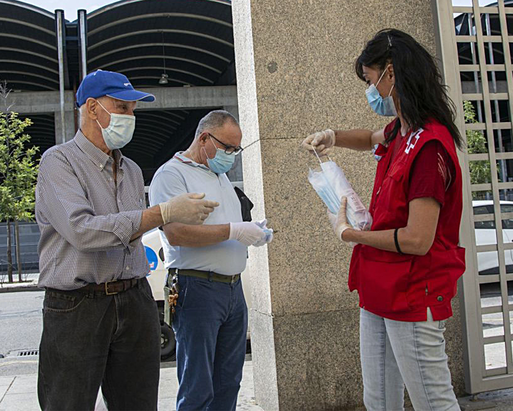Una voluntaria de Cruz Roja reparte mascarillas quirúrgicas a dos ciudadanos.
