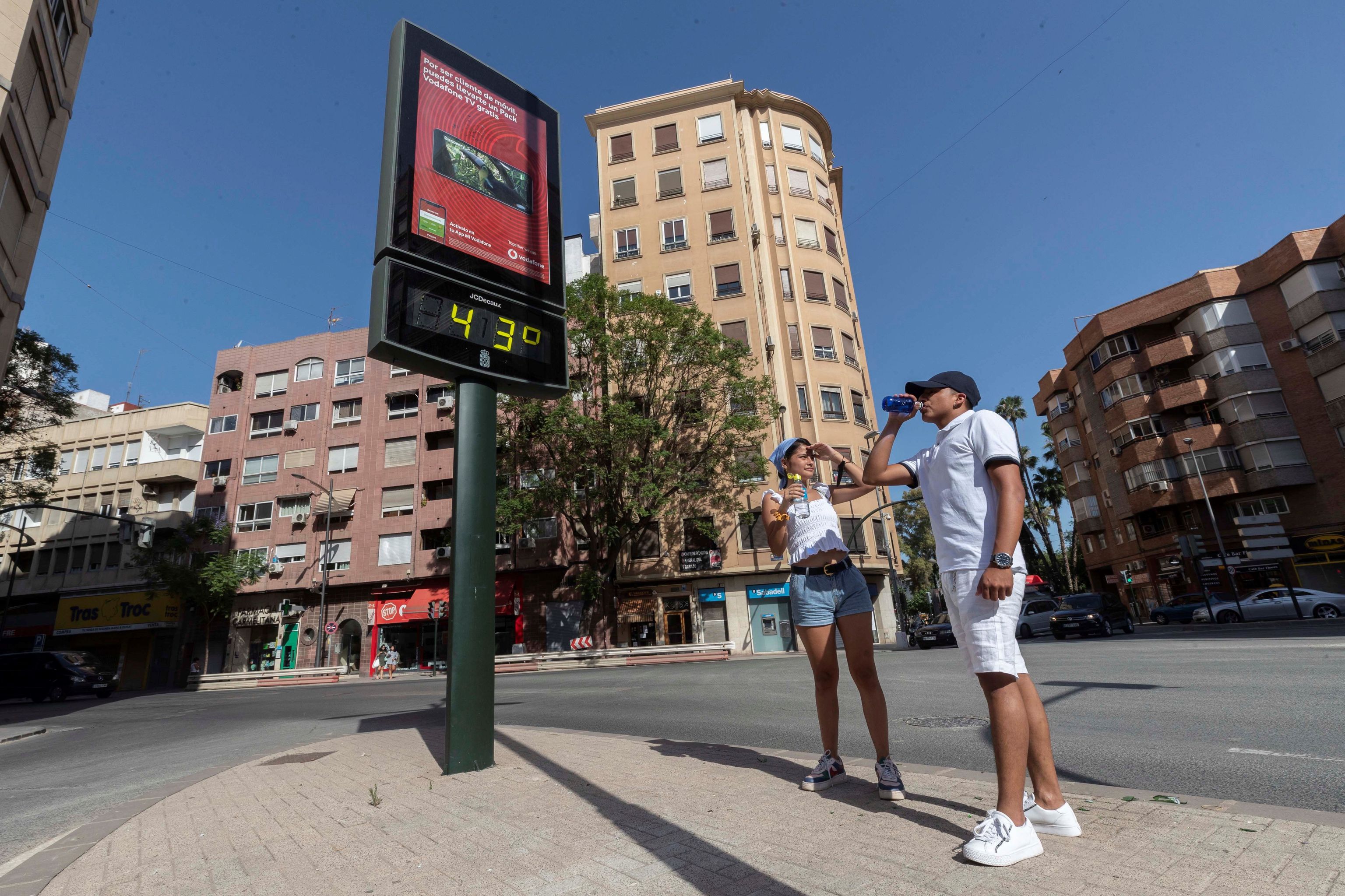Dos jóvenes beben agua junto a un termómetro que marca 43 grados centígrados, este martes en la Plaza del Royo de Murcia.