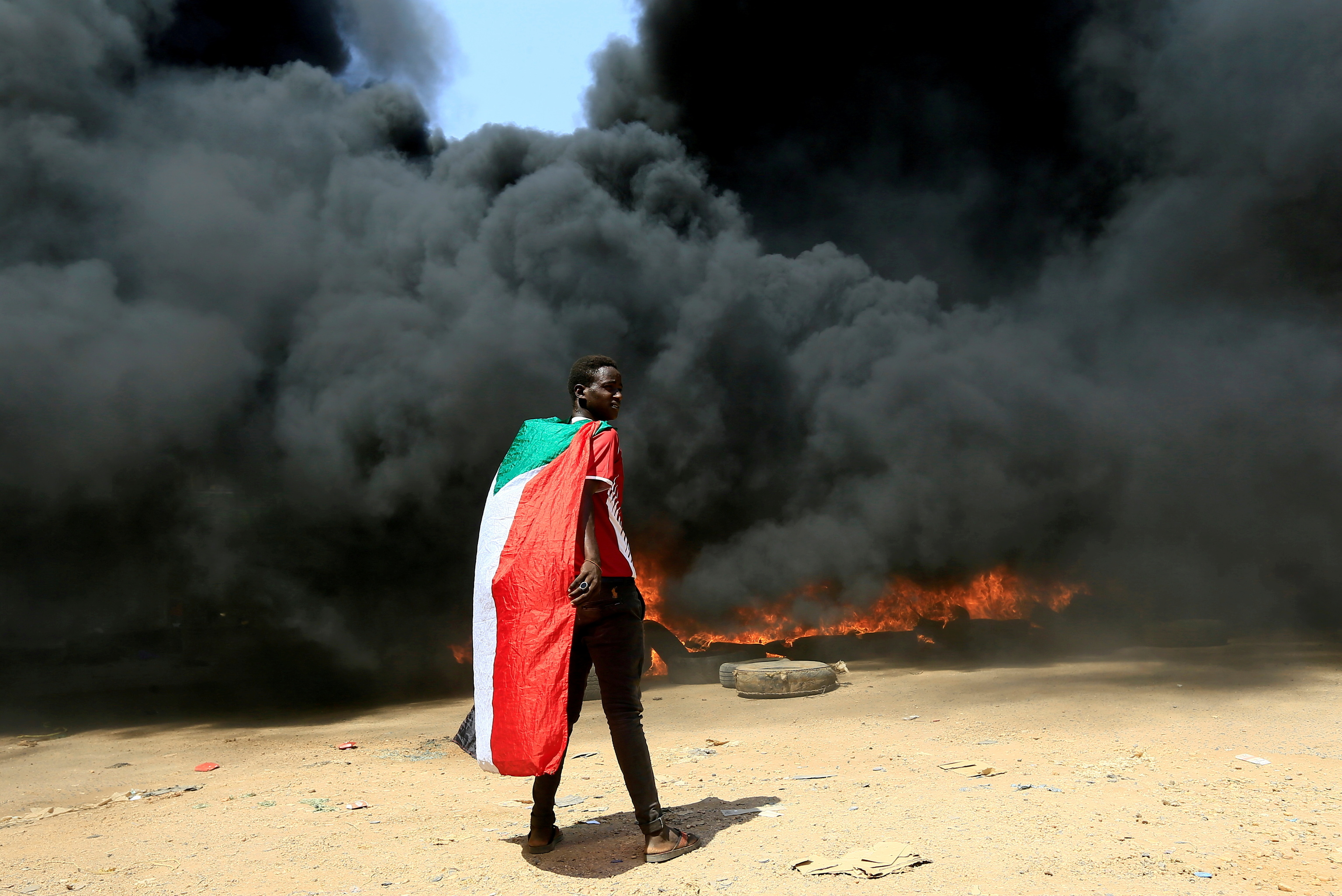 Una persona camina con la bandera de Sudn bajo una columna de humo durante una protesta en Jartum.