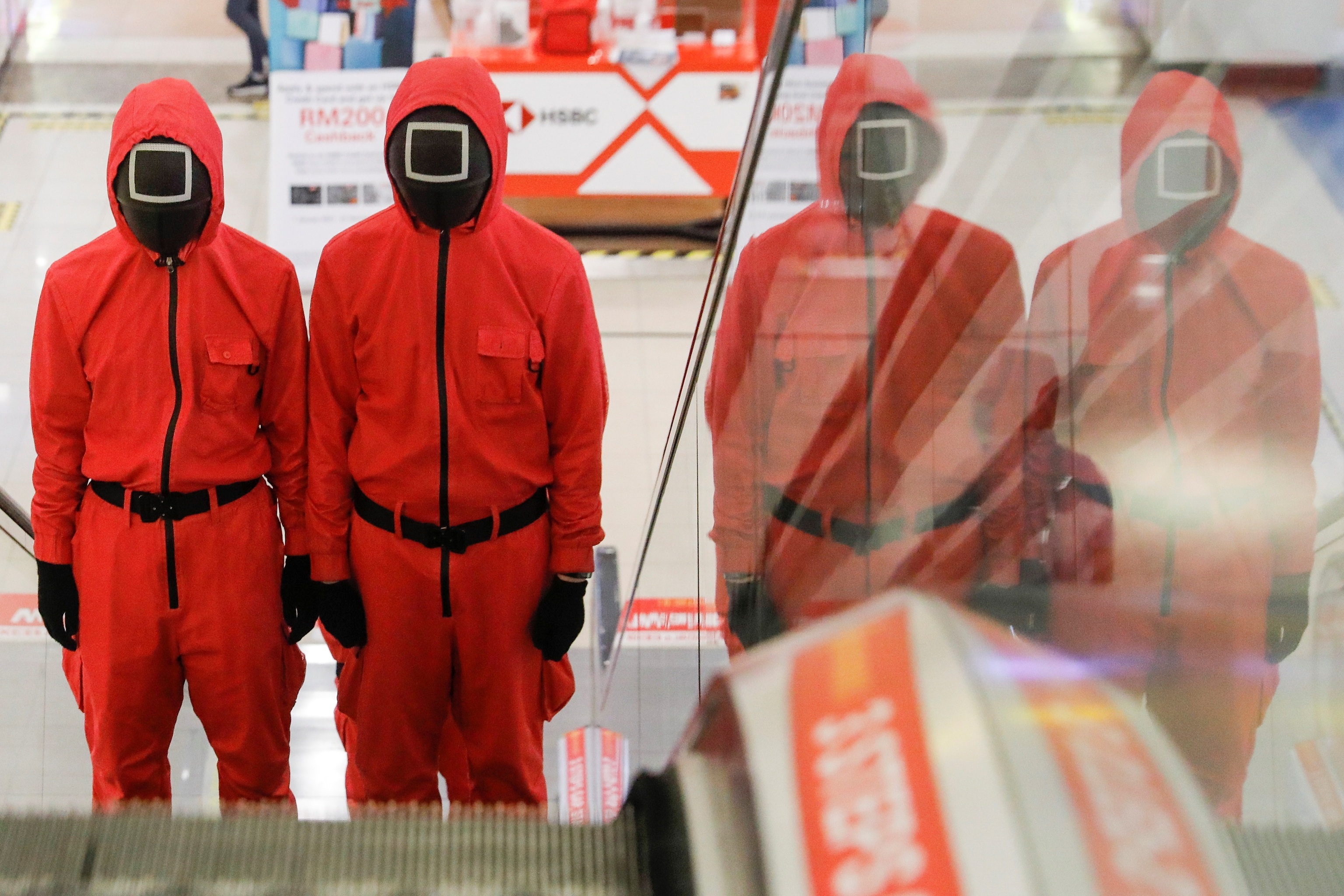 Dos personas disfrazadas de guardianes de la serie de Netflix El juego del calamar, subiendo las escaleras mecánicas de un centro comercial de Malasia.