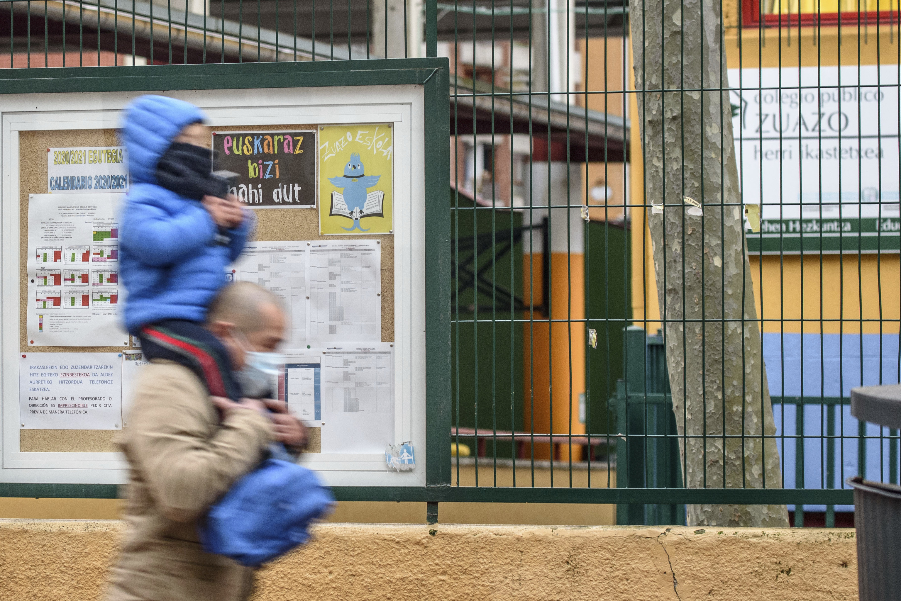 Un padre lleva en sus hombros a su hijo frente a un centro escolar con carteles en euskera.