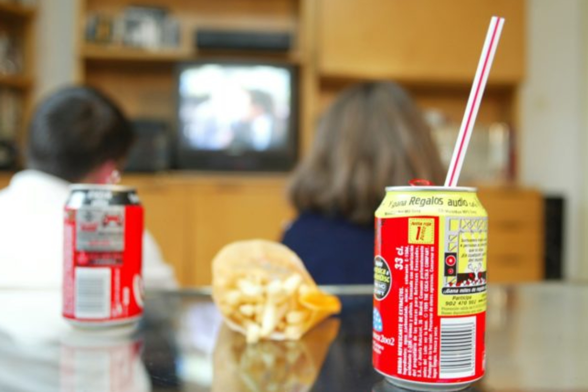 Dos niños tomando refrescos y patatas fritas mientras ven la televisión.