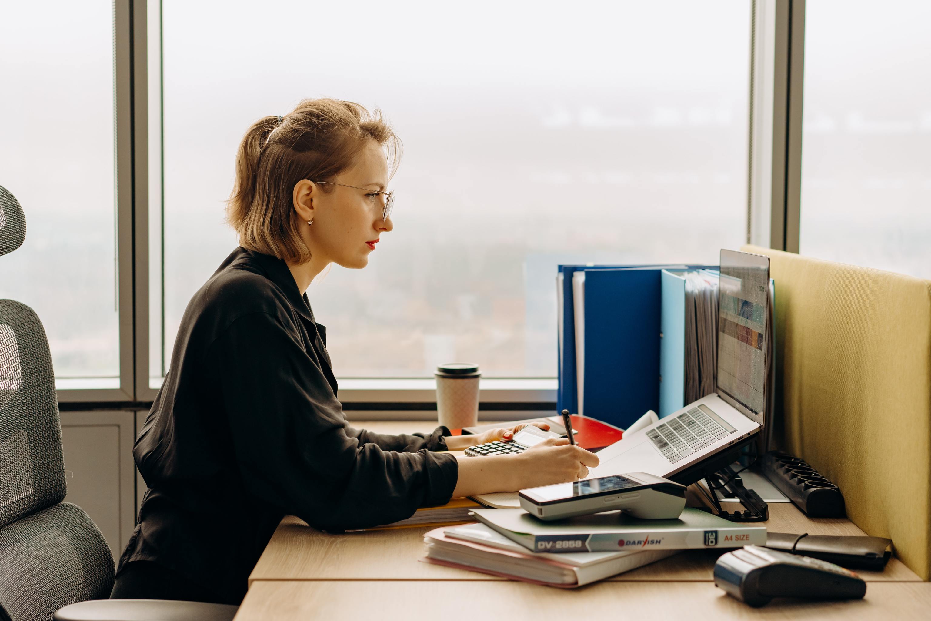 Una mujer trabaja con un ordenador, tomando apuntes y con una calculadora sobre la mesa.