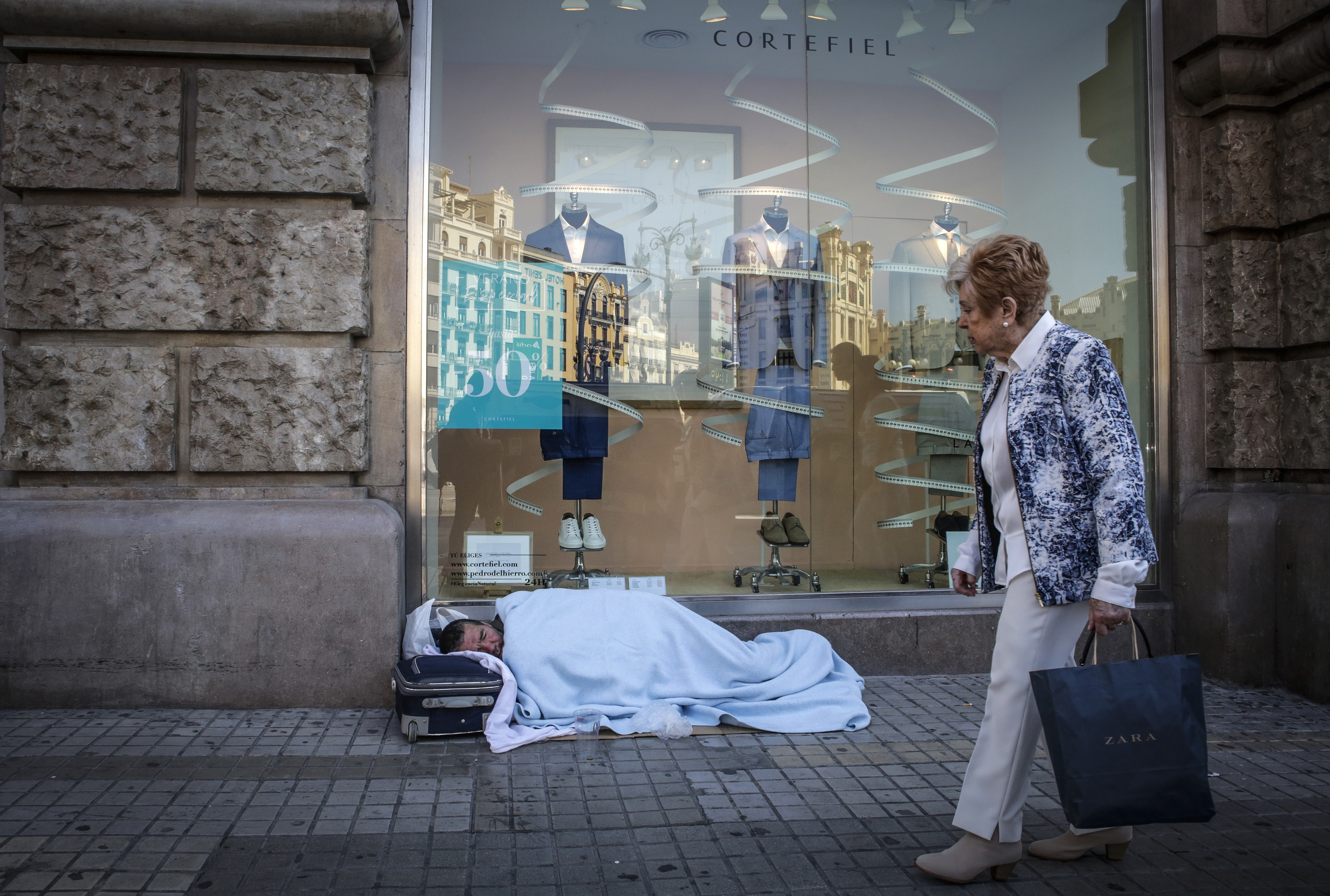 Una persona sin hogar duerme junto a un escaparate de una tienda, situada en una calle centrica de Valencia.
