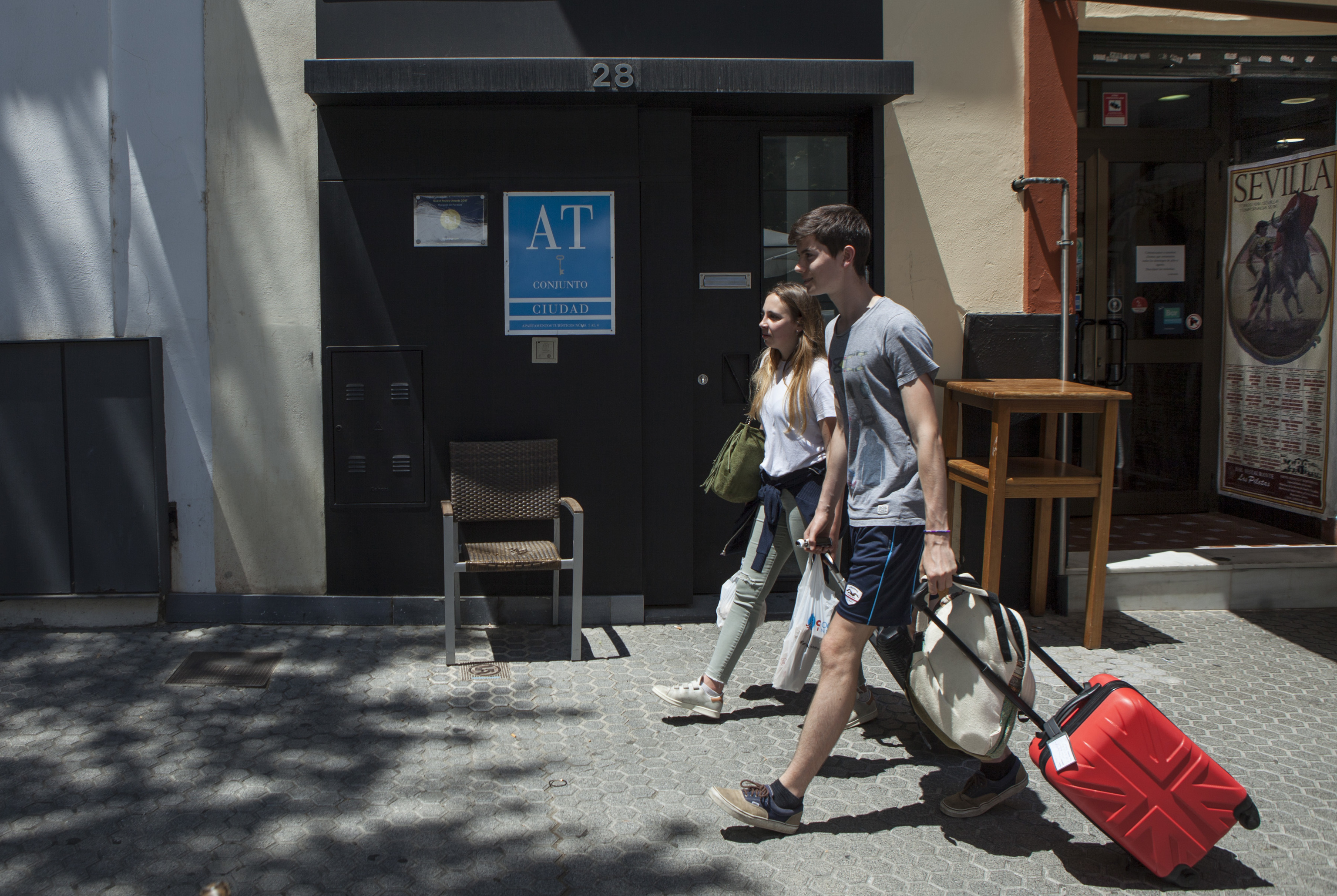 Dos turistas cargan con su maleta ante un alojamiento turstico.