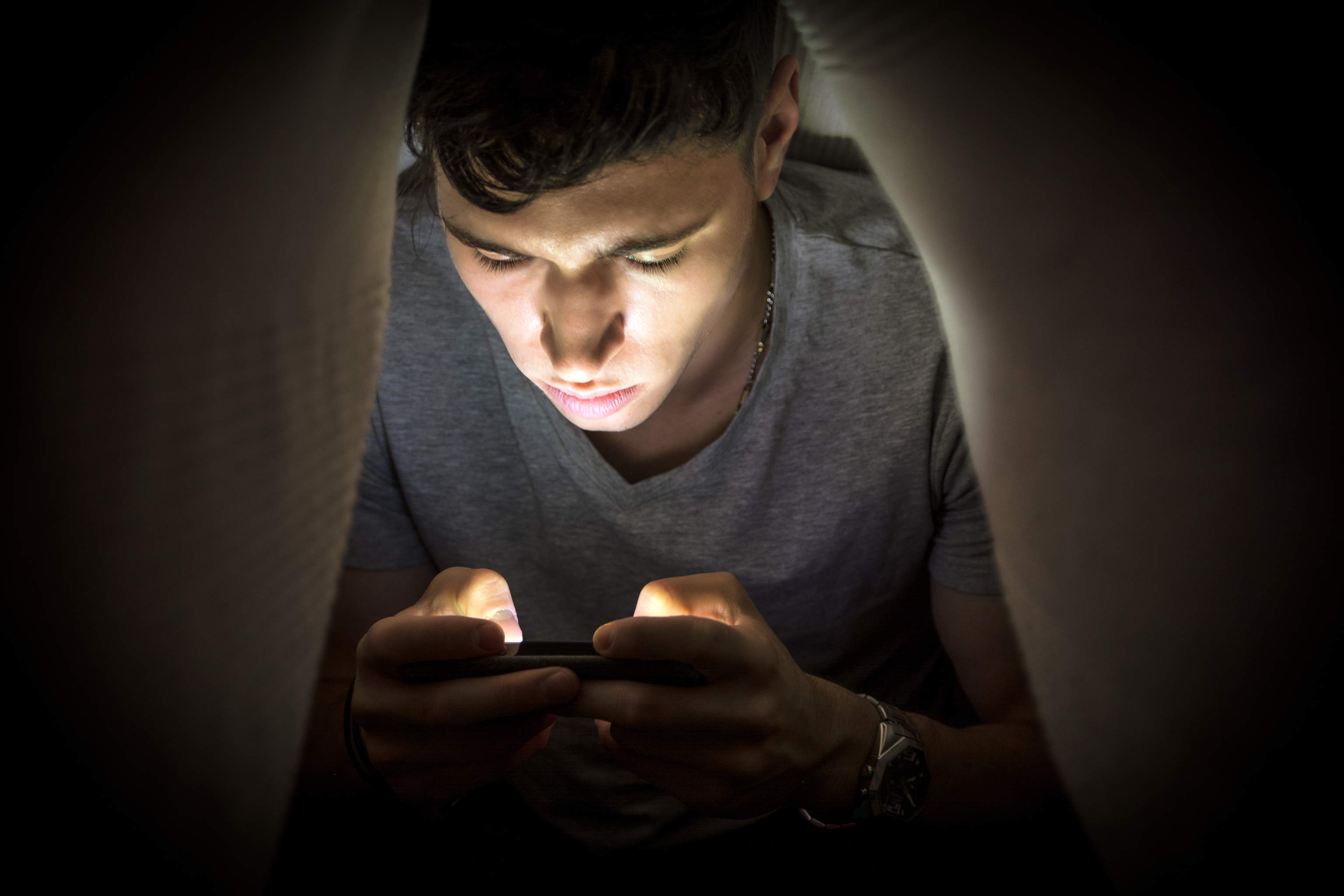 Un adolescente bajo una manta con un móvil.