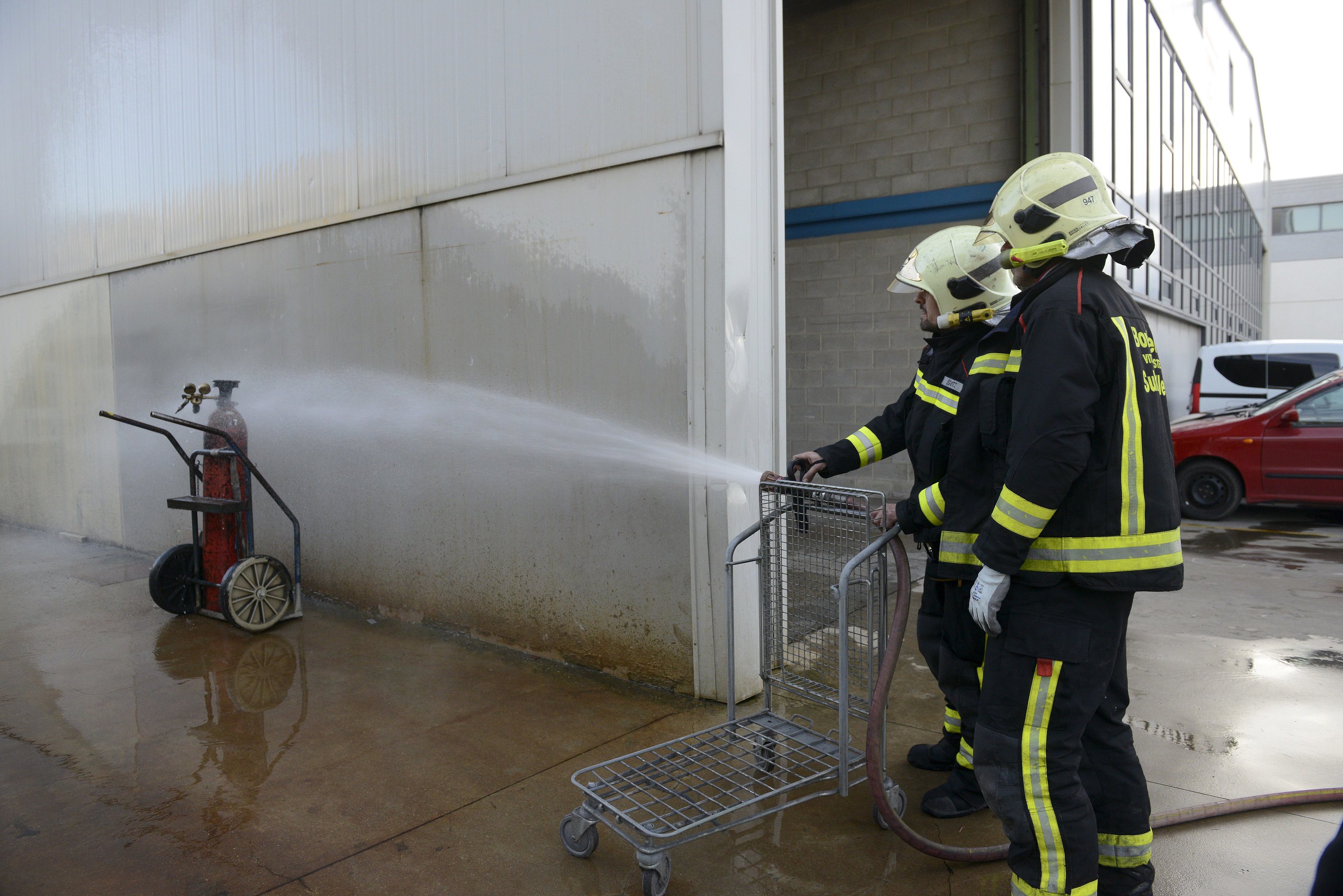 Dos bomberos realizan unas prcticas con una botella de gas en el exterior de un pabelln industrial.