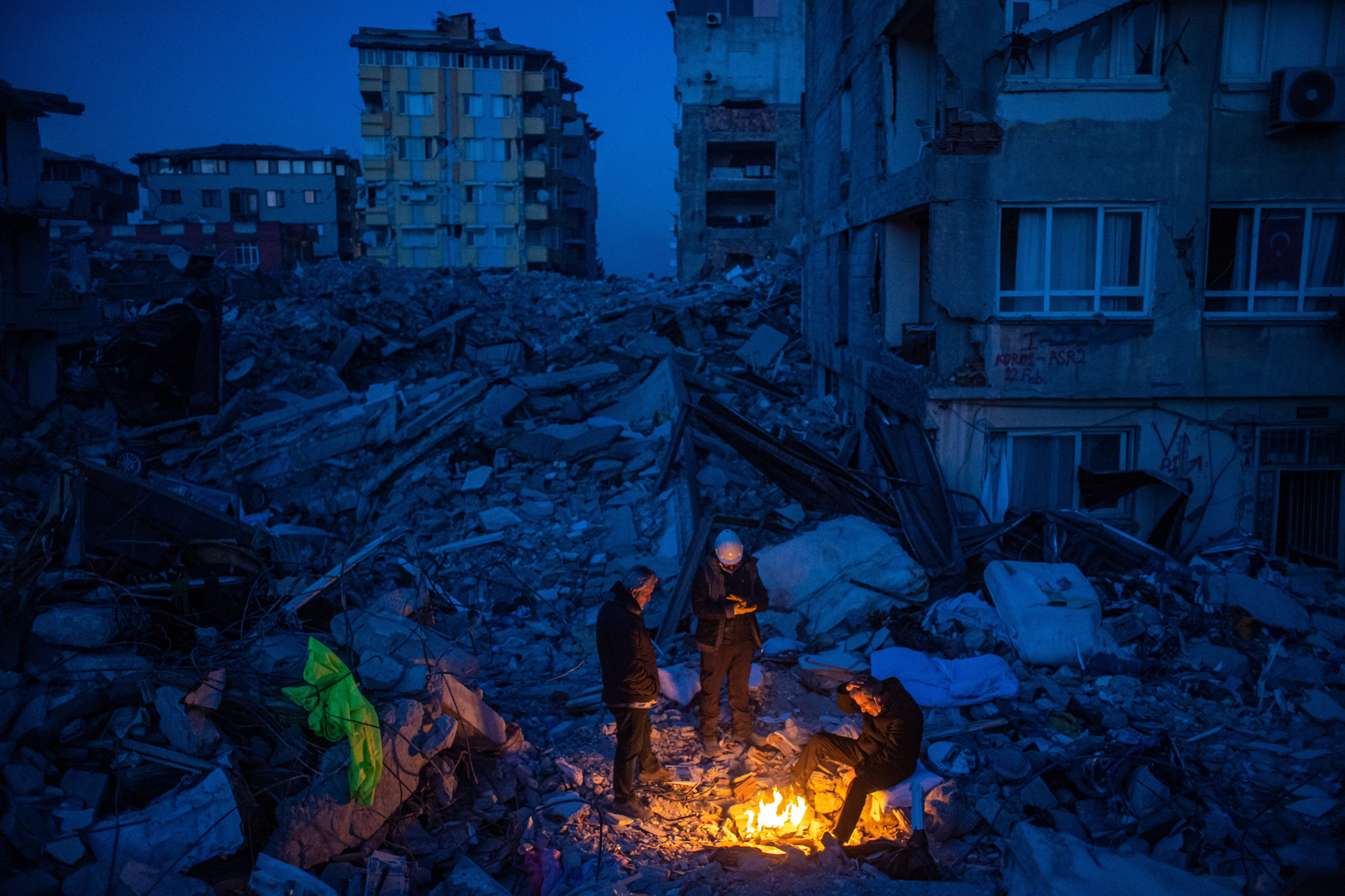 Personas afectadas por los sesmo de hace un ao en Turqua alrededor de una hoguera y entre escombros de edificios, en Hatay.